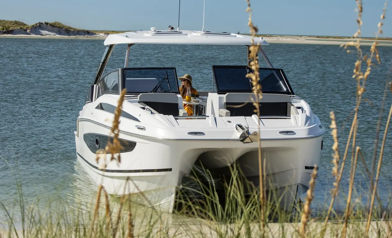 A modern white boat anchored in clear water, with a woman in a sunhat enjoying a drink, surrounded by tall grass.